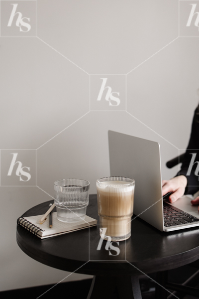 Modern workspace stock image featuring woman typing on laptop at desk with coffee, water, notebook and pen.