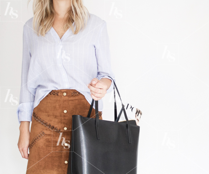 Woman in blue button up and brown skirt holding black leather purse.