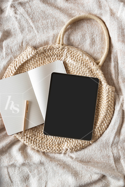 Styled stock photo featuring a blank iPad screen, notebook and wicker bag on bed, perfect for social media designs.