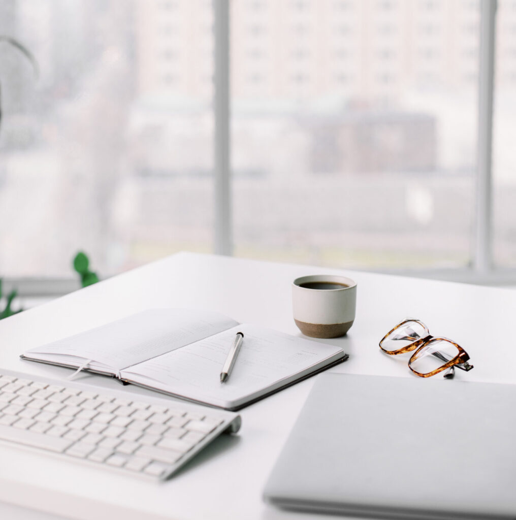 Urban scenery for minimal modern workspace featuring an open notebook next to a laptop and keyboard, coffee and reading glasses.