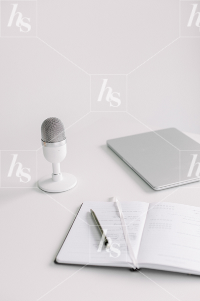 A styled stock photo of podcast microphone with notes and laptop beside it.