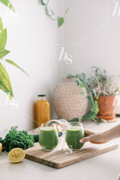 Stock photo of woman reaching for freshly prepared healthy green smoothie next to green plants and vegetables.
