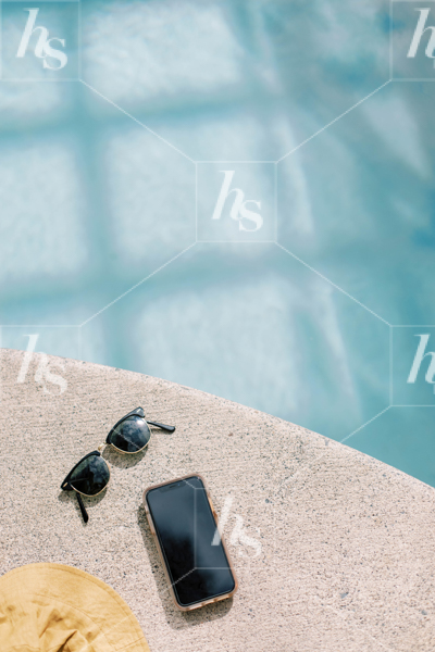 Flatlay of iPhone mockup on concrete next to sunglasses and hat on pool deck, perfect stock photo for your summer graphics.