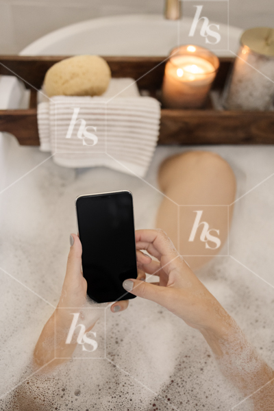 Woman scrolling phone while bathing in tub. Download this wellness and beauty collection from Haute Stock
