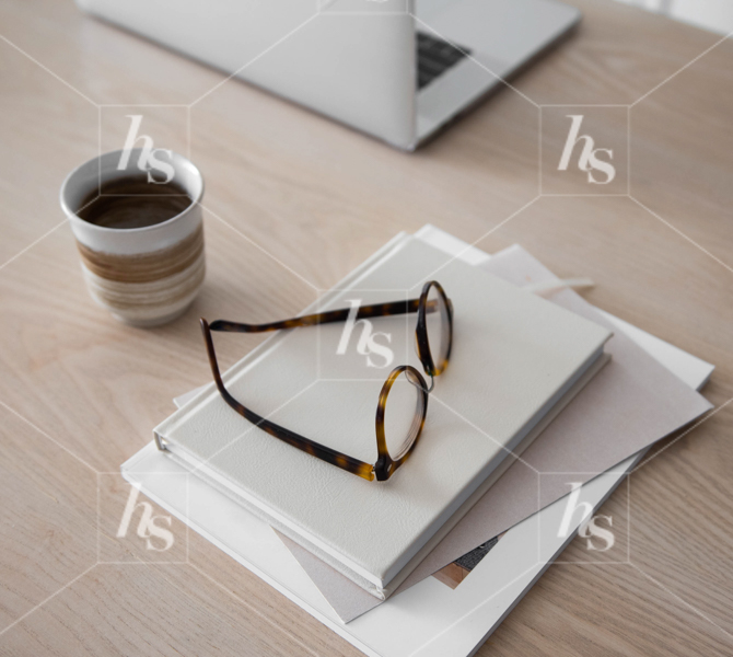 Reading glasses and stack of books next to laptop and coffee on desk, a peaceful interior and workspace image.