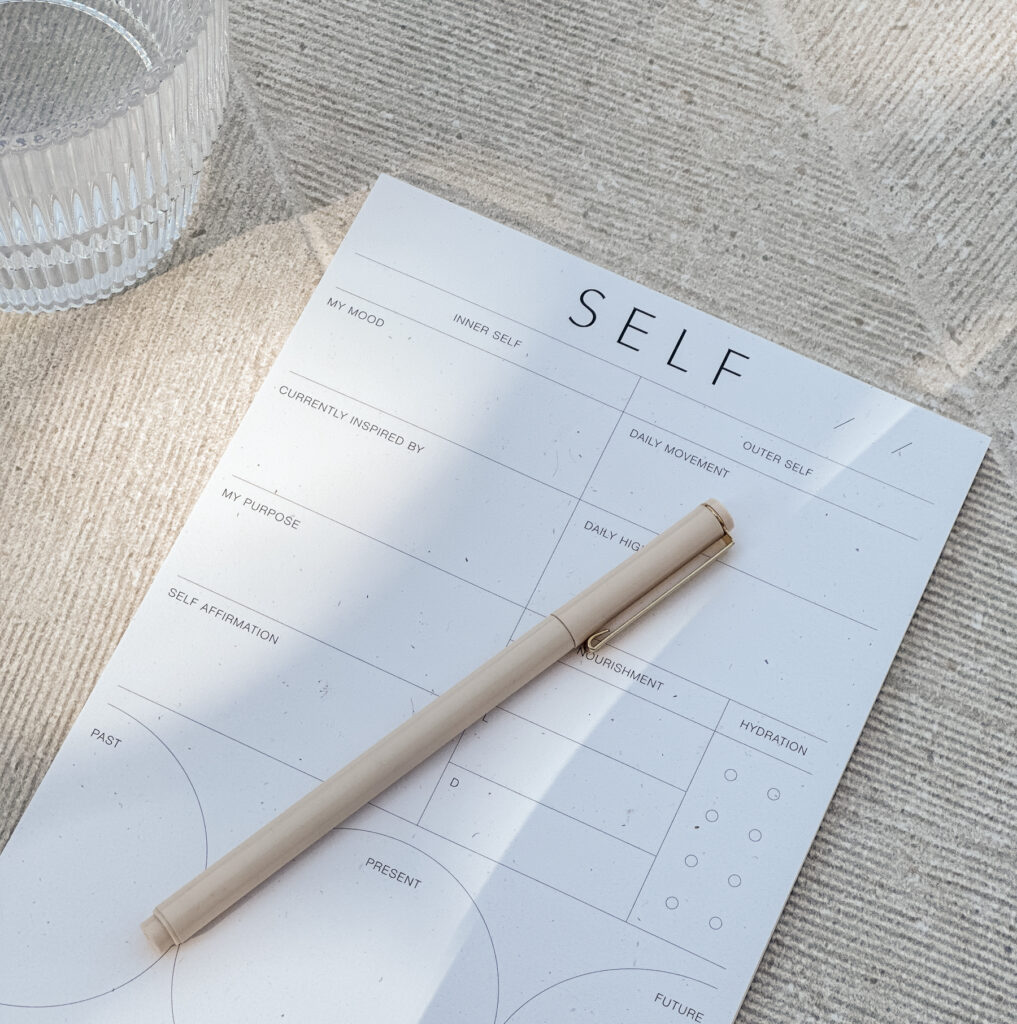 Neutral flatlay of a self care reflection worksheet labeled “SELF” with a beige pen and glass of water in soft sunlight.